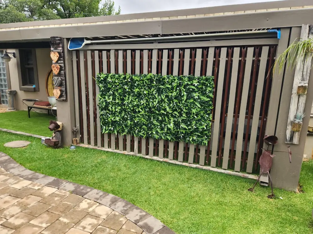 Foliage panel on timber slats: A rectangular foliage panel is centred on a timber-slat wall above artificial lawn and paving.